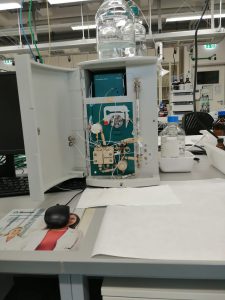 Ein geöffnetes technisches Gerät auf einem Tisch in einem Labor. Das Gerät zeigt interne Komponenten wie Schaltkreise und Kabel. Neben dem Gerät befinden sich ein Computermonitor, eine Maus und eine Wasserflasche. Auf dem Tisch liegt auch ein Blatt Papier.