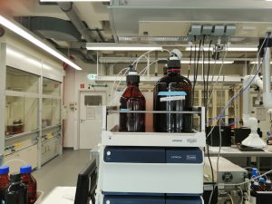 Laborumgebung mit mehreren Chemikalienflaschen auf einem Tisch. Im Vordergrund stehen zwei große braune Flaschen auf einem Laborgerät, während im Hintergrund Laboreinrichtungen und -geräte sichtbar sind.