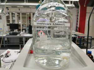 Ein großes Glasbehälter steht auf einem Tisch in einem Labor. Der Behälter enthält eine klare Flüssigkeit und ist mit einem Etikett beschriftet, das 