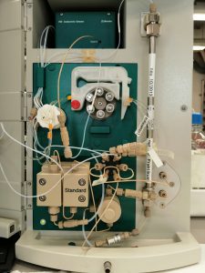 Technisches Gerät mit verschiedenen Rohren und Anschlüssen, das zur Durchführung von chemischen Analysen oder Experimenten verwendet wird. Die Oberfläche ist grün mit mehreren Knöpfen und einer Vielzahl von Verbindungen, die unterschiedliche Funktionen anzeigen.