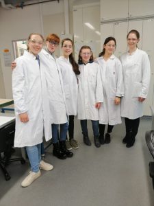 Sechs Personen in weißen Laborkitteln stehen lächelnd zusammen in einem Labor. Der Raum ist hell und modern eingerichtet, mit Arbeitsflächen im Hintergrund.