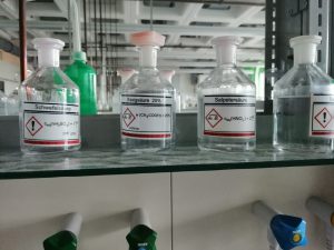 Vier Glasflaschen mit klarer Flüssigkeit stehen auf einem Regal. Jede Flasche hat ein Etikett mit chemischen Namen und Gefahrensymbolen. Im Hintergrund sind verschiedene andere Behälter und ein laborähnliches Umfeld sichtbar.