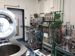 Laborumgebung mit verschiedenen wissenschaftlichen Geräten. Im Vordergrund befindet sich ein großes, rundes Behältnis, während im Hintergrund mehrere Maschinen und Instrumente mit Schläuchen und Anzeigen zu sehen sind. Eine Tür führt in einen angrenzenden Raum.
