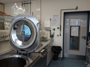 Eine moderne Laborumgebung mit einem großen, offenen Waschbecken und einer geschlossenen Tür im Hintergrund. Auf der linken Seite befindet sich ein großes, rundes Gerät, das wie ein Autoklav aussieht. An der Wand hängen verschiedene Utensilien und Anleitungen. Es gibt einen Mülleimer und eine kleine Gitterablage. Der Boden ist blau und die Wände sind hell.
