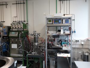 Laborumgebung mit verschiedenen Geräten: Auf der linken Seite steht ein komplexes Gerät mit mehreren Schläuchen und Anzeigen, während rechts ein Regal mit Monitoren, einem Messinstrument und weiteren Laborutensilien zu sehen ist. Die Wände sind mit Rohren und Kabeln ausgestattet, die zur technischen Infrastruktur gehören.