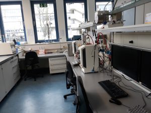 Ein moderner Laborraum mit zwei Arbeitsplätzen. Auf einem Tisch stehen zwei Computerbildschirme, ein Keyboard und verschiedene Geräte mit Kabeln. Im Hintergrund sind Regale mit weiteren Geräten und ein Fenster, durch das Tageslicht strömt. Der Boden ist blau und die Wände sind hell.