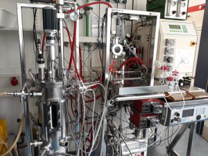 Ein komplexes Laborequipment mit verschiedenen Rohren, Schläuchen und Messgeräten. Die Apparatur besteht aus Edelstahl und ist mit mehreren Reglern sowie einem Bildschirm ausgestattet. Rote und transparente Schläuche verlaufen entlang der Struktur, und es sind verschiedene Instrumente zur Messung und Steuerung sichtbar.