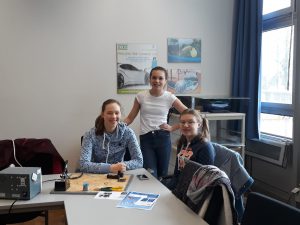 Drei Jugendliche sitzen und stehen an einem Tisch in einem Raum. Auf dem Tisch sind verschiedene Werkzeuge und Materialien zu sehen. Die Wände sind mit Postern dekoriert. Fenster lassen Tageslicht herein.