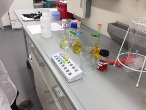 Laboratoriumstisch mit verschiedenen Reagenzgläsern und Flaschen, die unterschiedliche Flüssigkeiten in verschiedenen Farben enthalten. Im Hintergrund sind Laborausrüstung und ein Schreibtisch zu sehen.