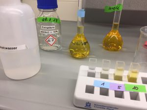 Laborumgebung mit verschiedenen Reagenzgläsern und Behältern. Ein transparenter Behälter mit Wasser, ein Etikett mit der Aufschrift 