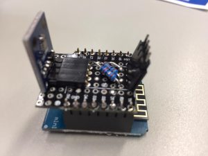 Ein elektronisches Bauteil auf einem Tisch, das mehrere Platinen und Pins zeigt. Die obere Platine hat einen kleinen Chip und einige elektronische Komponenten, während die untere Platine eine Basis mit Anschlüssen darstellt.