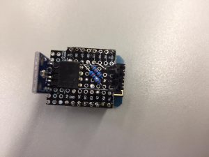 Ein Mikrocontroller-Board mit einer schwarzen Platine, das mit verschiedenen Komponenten und Löchern für Verbindungen ausgestattet ist. Es zeigt einen integrierten Schaltkreis in der Mitte und eine Anschlussstelle an der Seite. Das Bild ist von oben aufgenommen und zeigt die Details der Platine auf einem hellen Hintergrund.