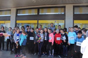 Eine Gruppe von Kindern und Jugendlichen steht in Sportkleidung vor einem Gebäude. Sie tragen Nummern auf ihren Shirts und lächeln in die Kamera. Im Hintergrund sind Fenster und eine gelbe Beschilderung sichtbar. Die Szene vermittelt eine fröhliche Atmosphäre, wahrscheinlich im Kontext eines sportlichen Events oder Wettkampfs.