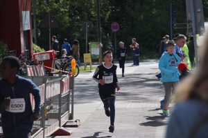 Ein junger Läufer in einem schwarzen Trikot läuft auf einer Straße, umgeben von Zuschauern und anderen Teilnehmern. Im Hintergrund sind Bäume und Verkehrszeichen sichtbar. Einige Personen stehen an der Seite, während andere sich ebenfalls in Bewegung befinden.