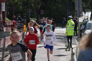 Eine Gruppe von Kindern läuft auf einer Straße während eines Rennens. Einige Kinder tragen Laufshirts mit Nummern, während ein Erwachsener in einer gelben Warnjacke am Rand steht. Im Hintergrund sind Fahrräder und andere Kinder zu sehen, die ebenfalls am Rennen teilnehmen.