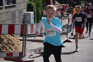 Ein Junge in einem blauen T-Shirt und schwarzen Hosen läuft mit entschlossenem Gesichtsausdruck in einem Wettlauf. Er trägt eine Nummer auf der Brust, während im Hintergrund weitere Kinder ebenfalls rennen. Baustellenabsperrungen sind sichtbar, und es ist ein sonniger Tag.
