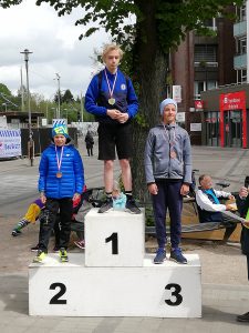 Drei Kinder stehen auf einem Siegerpodest. Das Kind in der Mitte trägt eine Medaille und ist in sportlicher Kleidung. Die beiden anderen Kinder stehen links und rechts von ihm, eines trägt eine blaue Jacke und das andere eine graue Jacke, beide haben ebenfalls Medaillen. Im Hintergrund sind Menschen und Gebäude zu sehen.