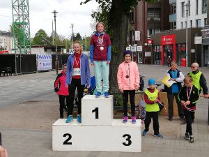 Drei Mädchen stehen auf einem Siegerpodest nach einem Wettkampf. Das Mädchen ganz oben trägt eine Medaille und hat einen stolzen Gesichtsausdruck. Die anderen beiden Mädchen stehen auf den zweiten und dritten Plätzen. Im Hintergrund sind Zuschauer und städtische Gebäude zu sehen.