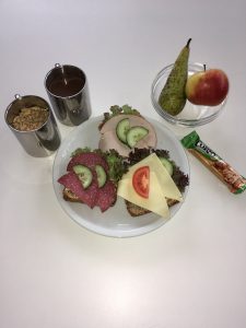 Ein Teller mit belegten Broten, darauf sind Salami, Schinken, Gurkenscheiben und Käse zu sehen, umgeben von frischem Salat. Daneben befinden sich zwei kleine Behälter mit Müsli und einem Getränk, sowie eine Birne und ein roter Apfel in einer Schale. Ein Snack-Riegel liegt ebenfalls auf dem Tisch.