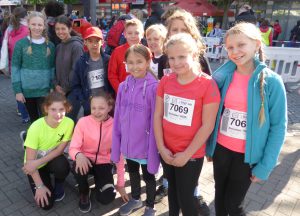 Gruppe von Kindern in Sportkleidung, die lächeln und für ein Foto posieren. Einige tragen Startnummern auf ihren Oberteilen. Im Hintergrund sind Stände und Menschen zu sehen, was auf eine Sportveranstaltung hinweist.