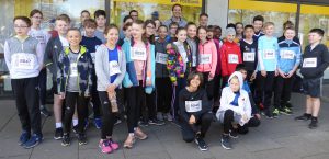 Gruppe von Kindern und Jugendlichen, die vor einem Gebäude stehen. Sie tragen Sportkleidung und haben Startnummern an ihren Oberteilen befestigt. Die Stimmung ist fröhlich und motiviert. Im Hintergrund sind große Fenster sichtbar.