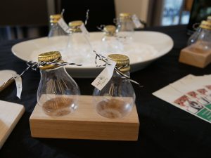 Zwei kleine Glasfläschchen mit goldenen Deckeln stehen auf einem Holzständer. Im Hintergrund sind weitere Fläschchen auf einem weißen Teller und einige Karten zu sehen. Der Tisch ist mit einem schwarzen Tuch bedeckt.