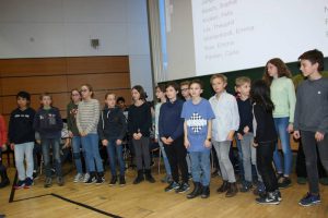 Eine Gruppe von Kindern und Jugendlichen steht auf einer Bühne in einem Saal. Sie sind in verschiedenen Kleidungsstilen gekleidet und scheinen auf einen Auftritt oder eine Präsentation vorbereitet zu sein. Im Hintergrund ist eine Projektionsfläche sichtbar, auf der Texte oder Informationen angezeigt werden. Der Raum hat eine helle Holzoptik und eine freundliche Atmosphäre.