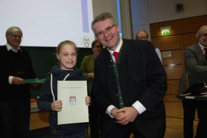 Ein lächelndes Mädchen hält ein Zertifikat in der Hand und steht neben einem Mann in Anzug mit Krawatte. Im Hintergrund sind weitere Personen zu sehen. Die Szene findet in einem Raum mit Präsentationstechnik statt.