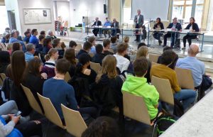 Eine Gruppe von Menschen sitzt in einem Raum und hört einer Diskussionsrunde zu. Auf der Bühne stehen mehrere Redner hinter Tischen. Das Publikum, bestehend aus jungen Erwachsenen und Jugendlichen, blickt in Richtung der Redner. Der Raum ist hell erleuchtet und modern eingerichtet.