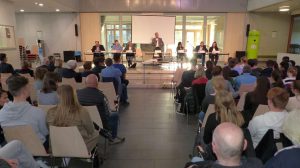 Eine Versammlung in einem großen Raum, bei der mehrere Personen auf einer Bühne sitzen und sprechen. Das Publikum besteht aus vielen Zuhörern, die in Reihen von Stühlen sitzen. Im Hintergrund ist eine Leinwand sichtbar.