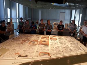 Eine Gruppe von Kindern und Jugendlichen steht um einen großen, detaillierten Stadtplan oder eine Karte in einem Museum. Die Karte ist auf einem Tisch ausgebreitet, und die Besucher schauen interessiert darauf. Im Hintergrund sind helle Fenster und weitere Ausstellungsstücke zu sehen.