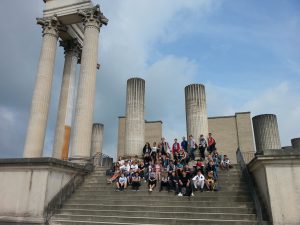 Eine große Gruppe von Menschen sitzt auf den Stufen vor einer historischen Säulenarchitektur. Die Säulen sind teilweise beschädigt und stehen hinter den Personen. Der Himmel ist bewölkt, und die Szene vermittelt einen Eindruck von Freizeit und Erkundung.