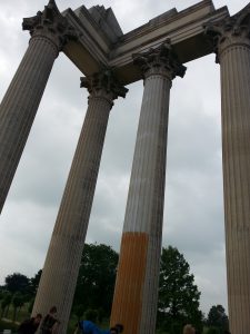 Ein Bild von mehreren hohen, klassischen Säulen mit Kapitellen, die in einem parkähnlichen Bereich stehen. Einige Säulen sind beschädigt oder unvollständig, wobei eine Säule teilweise orange gestrichen ist. Der Himmel ist bewölkt, und im Hintergrund sind Bäume sichtbar.