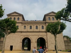 Eine historische Architektur mit zwei Türmen und großen Bögen im Erdgeschoss. Vor dem Gebäude stehen einige Menschen, während Bäume mit runden Kronen die Szene umgeben. Der Himmel ist bewölkt, was eine ruhige Atmosphäre schafft.