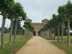 Ein gepflasterter Weg führt durch eine Allee von Bäumen zu einem historischen Gebäude mit zwei Türmen und einem großen Tor. Der Himmel ist bewölkt, und die Umgebung ist grün und gut gepflegt.