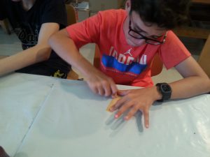 Ein junger Mensch mit Brille und einem roten T-Shirt arbeitet konzentriert an einem Holzstück auf einem Tisch. Seine linke Hand hält das Holz, während er mit der rechten Hand ein Werkzeug benutzt. Im Hintergrund sind weitere Personen zu sehen, die ebenfalls an einem Tisch sitzen.