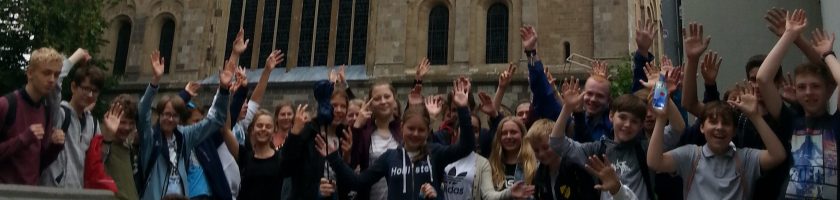 Gruppenszene vor einem historischen Gebäude, in der viele Personen lächeln und die Hände in die Luft heben, um Freude oder Begeisterung auszudrücken.