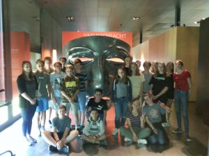 Gruppe von Jugendlichen posiert vor einer großen, schwarzen Maske in einem Museum. Im Hintergrund ist eine rote Wand mit dem Wort 
