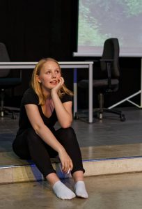 Eine junge Frau mit blonden Haaren sitzt auf dem Boden und schaut nachdenklich. Sie trägt ein schwarzes Oberteil und schwarze Leggings, sowie weiße Socken. Im Hintergrund sind Büromöbel und eine Projektion sichtbar.