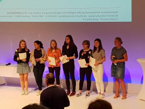Sechs junge Frauen stehen auf einer Bühne, halten Urkunden oder Dokumente in den Händen und lächeln. Im Hintergrund ist eine Präsentation auf einem Bildschirm sichtbar. Die Szene scheint eine Auszeichnung oder Ehrung zu zeigen.