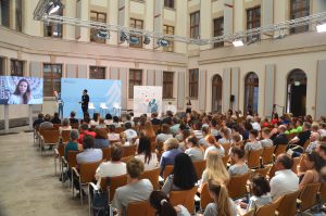 Eine große Gruppe von Menschen sitzt in einem Saal und schaut auf eine Präsentation. Im Vordergrund stehen zwei Personen, eine davon spricht, während die andere auf einen Bildschirm schaut, der eine Videopräsentation zeigt. Der Raum hat hohe Decken und große Fenster, mit einer Vielzahl von Stühlen, die in Reihen angeordnet sind.