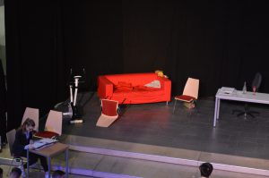 Bühnenbild mit rotem Sofa, zwei Stühlen und einem Tisch. Auf dem Sofa liegt ein Kissen, daneben steht ein Fahrrad. Im Vordergrund sitzt eine Person am Tisch und liest. Der Hintergrund ist schwarz.