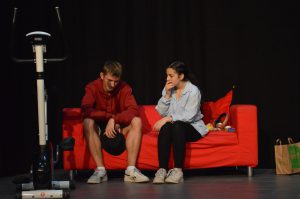 Zwei junge Menschen sitzen auf einem roten Sofa. Der Junge trägt ein rotes Sweatshirt und Shorts, während das Mädchen eine helle Bluse und schwarze Hose trägt. Beide wirken nachdenklich. Neben dem Sofa steht ein Fitnessgerät. Im Hintergrund ist eine dunkle Wand zu sehen. Rechts neben dem Sofa steht eine Papiertüte.