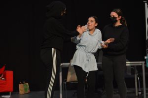 Drei Schauspielerinnen auf einer Bühne in einem dramatischen Moment. Eine der Frauen trägt ein schwarzes Outfit und steht mit erhobenen Händen da, während eine andere in einem hellen Oberteil und mit einem besorgten Gesichtsausdruck dazwischen steht. Die dritte Schauspielerin, ebenfalls in Schwarz, beobachtet die Szene. Im Hintergrund sind ein Tisch und einige Requisiten zu sehen.
