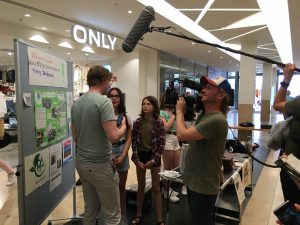 Drei Personen stehen vor einem Informationsstand in einem Einkaufszentrum. Eine Person hält ein Mikrofon über eine andere Person, die spricht, während eine dritte Person mit einer Kamera filmt. Im Hintergrund ist ein Schild mit der Aufschrift 