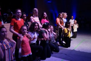 Eine Gruppe von jungen Frauen und Mädchen steht und kniet auf einer Bühne. Sie tragen bunte T-Shirts und posieren fröhlich für das Bild. Im Hintergrund sind schwaches Licht und ein Publikum zu erkennen.