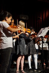 Eine Gruppe von Kindern spielt gemeinsam Geigen auf einer Bühne. Einige Kinder tragen weiße T-Shirts, während andere Kleider in verschiedenen Mustern anhaben. Im Hintergrund sind Notenständer und eine dunkle Bühnenumgebung mit roten Vorhängen zu sehen.