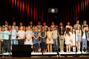 Eine Gruppe von Kindern steht auf einer Bühne und singt. Sie sind in mehreren Reihen angeordnet und tragen verschiedene bunte Kleidungsstücke. Im Hintergrund ist ein dunkler Vorhang mit rotem Licht. Einige Mikrofone stehen auf der Bühne.