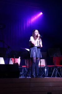 Eine Sängerin steht auf der Bühne und hält ein Mikrofon in der Hand. Sie trägt ein weißes Oberteil und einen schwarzen Rock. Im Hintergrund sind Stühle und Musikständer zu sehen, während lila Lichtstrahlen die Szene beleuchten.