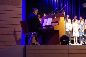 Ein Musiker sitzt an einem Klavier und spielt. Im Hintergrund steht ein Kinderchor, der aus mehreren Kindern besteht. Die Szene ist mit blauer Beleuchtung gestaltet und zeigt eine Aufführung auf einer Bühne.